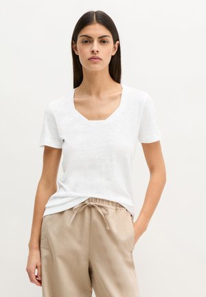 Femme aux cheveux lisses et foncés, portant un t-shirt blanc à manches courtes avec col en V et un pantalon beige à cordon, debout avec une main dans la poche.