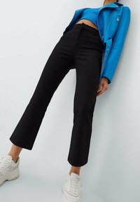 Pantalons évasés noirs avec une texture lisse, associés à un haut cropped bleu et une veste en cuir synthétique bleue, portés avec des baskets blanches.