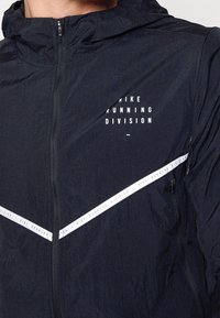 Gros plan d'une personne portant une veste noire Nike Running Division avec des rayures diagonales blanches et une fermeture éclair frontale.