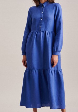 Kobieta w niebieskiej sukience midi z długim rękawem, zapinanej na guziki, z warstwowym dołem i dopasowanymi mankietami, stojąca na neutralnym tle.