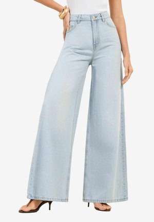 Nő világoskék magasderekú, széles szárú farmernadrágban, fehér felsőben, arany karpereccsel és fekete, nyitott orrú magassarkúban, egyik kezét a csípőjén tartva áll.