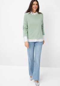 Lichtgroene textuurtrui gecombineerd met een wit shirt, blauwe flared jeans en zilveren spitse schoenen. De outfit is casual en gelaagd.