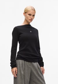 Schwarzer Strickpullover mit Rundhalsausschnitt und langen Ärmeln. Besitzt ein kleines gesticktes Logo auf der Brust. Kombiniert mit grauen, plissierten Hosen.