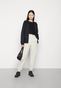 Schwarze Zip-Hoodie, schwarzes, kurzes Shirt, cremefarbene Sweatpants, schwarze Sneakers und eine kleine schwarze Handtasche. Glatte Texturen und lockere Passform.