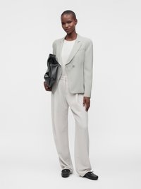 Femme portant un blazer gris clair, un haut blanc, un pantalon large blanc, des chaussures noires, tenant une grande pochette noire pliée, debout sur fond blanc.