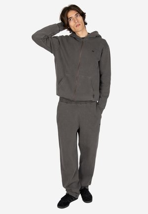 Junger Mann trägt dunkelgrauen Zip-Hoodie und passende Hose, steht mit einer Hand hinter dem Kopf und der anderen in der Tasche.