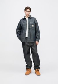 Ciemna kurtka dżinsowa z dużymi przednimi kieszeniami, zapinana na guziki, z kontrastującymi przeszyciami. Do tego luźne ciemne dżinsy i beżowe buty.