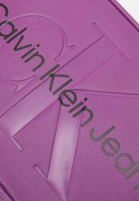 Superficie texturizzata viola con un grande logo in rilievo "CK" e il testo stampato in nero "Calvin Klein Jeans" sopra di esso.