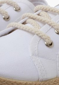 Adidași albi din pânză cu șireturi împletite bej, ochiuri argintii și o talpă texturată asemănătoare cu paiele. Prezintă accente cusute pentru durabilitate.