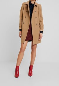Cappotto doppiopetto color cammello con bottoni argentati, abbinato a una gonna rossa a fantasia e stivaletti rossi con lati neri.