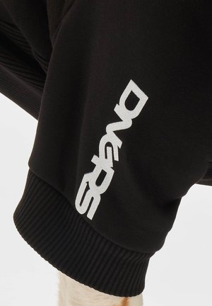 Mustat hupparit, joissa on ribbaus. Vasemmalla puolella on rohkealla valkoisella tekstillä, joka lukee "DIVERS." Materiaali näyttää pehmeältä ja siinä on tekstuuri.