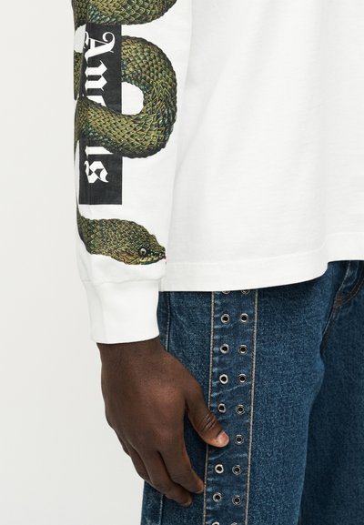 Λευκό πουκάμισο με μακριά μανίκια που διαθέτει πράσινο σχέδιο φιδιού στο μανίκι μαζί με έντονο κείμενο. Συνδυάζεται με μπλε τζιν παντελόνι με μεταλλικά καρφιά.