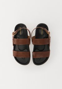Sandalias de cuero marrón con dos tiras anchas y correa ajustable en el tobillo, que cuentan con una suela texturizada negra y el nombre de la marca en relieve.
