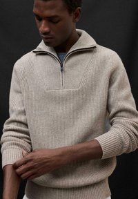 Beiger Strickpullover mit einem Reißverschlusskragen, gerippten Ärmelbündchen und strukturiertem Muster. Modell passt den Ärmel an und zeigt die Webart des Stoffes.