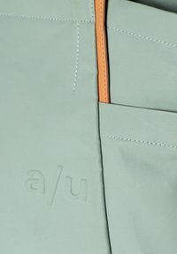 Lichtgroene tas met een gestructureerd oppervlak, voorzien van oranje accenten en een zijvak. Geëmbosseerd logo "a/u" op het materiaal.