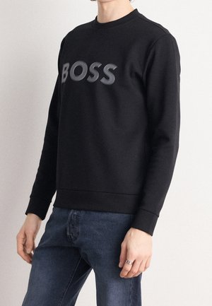 Homme portant un sweat-shirt noir avec le texte "BOSS" à l’avant, associé à un jean bleu, debout devant un fond uni.