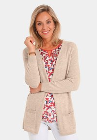 Beige, gebreide cardigan met open voorkant, zijzakken en geribbelde manchetten, gelaagd over een rood en blauw patroon top.