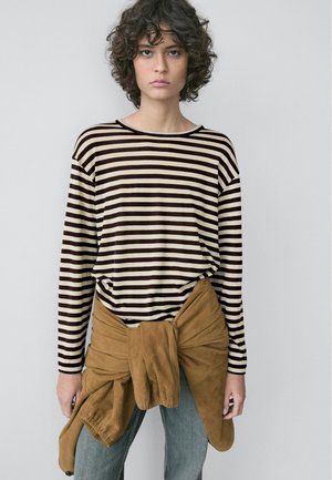 Junge Person mit lockigem Haar, die ein schwarz-weiß gestreiftes Langarmshirt trägt, eine braune Jacke um die Taille gebunden hat und blaue Jeans anhat.