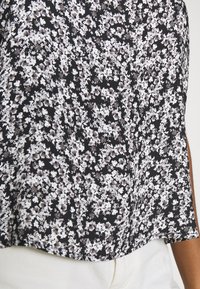 Tecido preto com um padrão floral em tons de branco e cinza, com uma textura suave e um corte descontraído, usado sobre calças brancas.