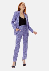Purple patterned suit with a fitted blazer and matching trousers, featuring a vibrant design and textured fabric. Black top underneath.