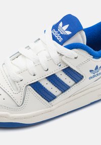 White leather sneaker featuring blue accents, three blue stripes, white laces, and a blue padded tongue with branding. Textured sole.