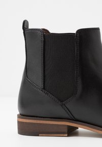 Černá kožená kotníková bota s pružnými bočními panely, kulatou špičkou a hnědým naskládaným podpatkem s horizontálními pruhy. Hladký povrch.