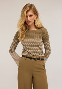 Top tricotat cu model în zigzag în tonuri pământii de verde și bej, având un guler rotund și mâneci lungi; asortat cu pantaloni bej cu talie înaltă.