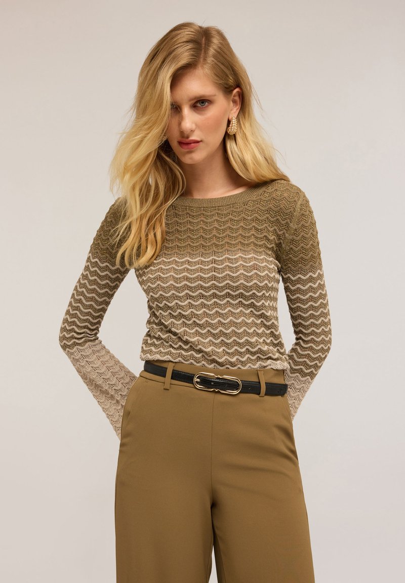 Top tricotat cu model în zigzag în tonuri pământii de verde și bej, având un guler rotund și mâneci lungi; asortat cu pantaloni bej cu talie înaltă.