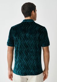Koszulka z krótkim rękawem w kolorze teal z welurowego materiału, z uniesionym falistym wzorem. Tył ma gładką teksturę i kołnierzyk.