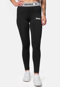 Black leggings featuring a stretchy, smooth fabric, a white branded waistband, and a small logo on the thigh. Designed for active wear.