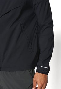 Czarna kurtka z długim rękawem, zapinana na zamek, z boczną kieszenią, noszona przez osobę, z odblaskowym paskiem na mankiecie rękawa oraz ciemnosiwymi spodniami.