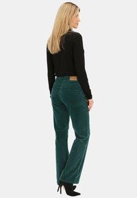 Turkusowe spodnie z korduroju o prostym kroju, z brązową skórzaną łatą w talii oraz detalem w postaci tylnej kieszeni. W połączeniu z czarną bluzką.