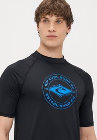Svart rashguard med kort ärm och en blå Rip Curl-logotyp i cirkulär design, med mjukt, stretchigt tyg och platta sömmar.