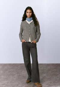 Una mujer con cabello largo y oscuro viste un cárdigan gris, una bufanda azul al cuello, jeans de pierna ancha negros y zapatos mocasín marrones, de pie sobre un suelo gris.
