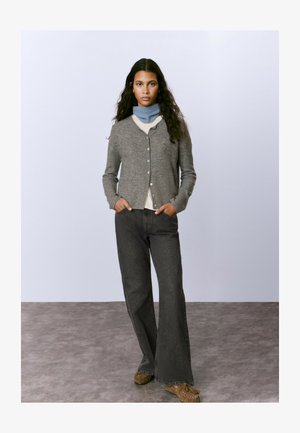 Una mujer con cabello largo y oscuro viste un cárdigan gris, una bufanda azul al cuello, jeans de pierna ancha negros y zapatos mocasín marrones, de pie sobre un suelo gris.