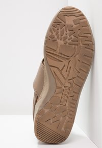 Suela de zapato marrón claro con caucho texturizado, que presenta un patrón de hojas y un logo. El lateral tiene un acabado liso y una ligera curva.