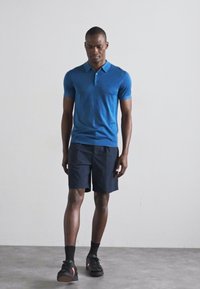 Sinine polo särk lühikeste varrukatega, koos tumehallide lühikeste pükstega. Mudel kannab musti tossusid ja lühikesi sokke. Lihtne, puhas disain.