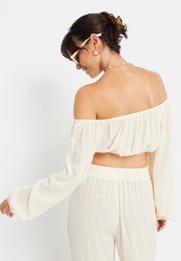 Cremfarbener, schulterfreier Crop-Top mit langen Ärmeln und strukturiertem Stoff. Verfügt über einen elastischen Taillenbund und wird mit passenden Hosen kombiniert.