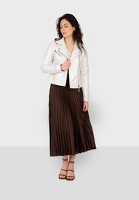 Bijela kožna motoristička jakna, naborana smeđa maksi suknja i smeđi sandali s remenčićima. Kosa je stilizirana slobodno, a osoba se nalazi protiv neutralne pozadine.