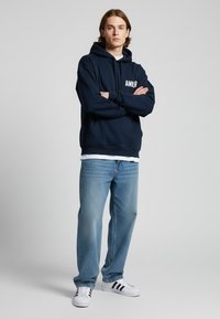 Felpa navy con stampa "AMLR", in misto cotone, vestibilità ampia, abbinata a jeans azzurri chiari e scarpe da ginnastica bianche con strisce nere.