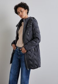 Mörkgrå, vadderad pufferjacka med huva, dragkedja fram och mockakänsla. Har silverfärgad hårdvara och sidofickor. Kombinerad med blå jeans.