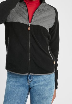 Czarna polarowa kurtka z pikowanymi szarymi panelami na ramionach, noszona na czerwonym golfie, w połączeniu z niebieskimi dżinsami.