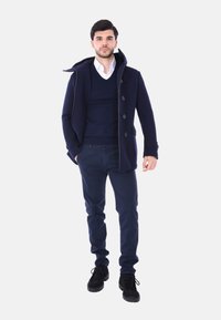 Giacca esterna blu con cappuccio, abbottonatura frontale e tasche laterali, indossata sopra un maglione blu e una camicia bianca, abbinata a pantaloni scuri e scarpe nere.