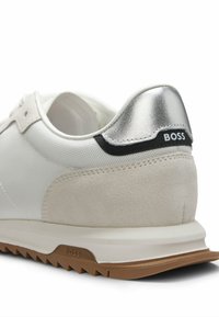 Valkoinen tennari, jossa on teksturoitu verkkopäällinen, mokkanahkaiset yksityiskohdat, metallinhohtoinen hopeanvärinen kantapään lappu sekä kumipohja ja kuminainen ulkopohja.