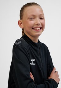 Svart zip-up jacka med meshstruktur, märkt med en vit logotyp. Har mönstrade grå accenter på axeln och hög krage.