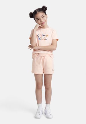 Mladé dievča s rozpustenými vlasmi v dvoch uzlíkoch, oblečené v broskyňovom tričku Calvin Klein a zodpovedajúcich šortkách, so šedými ponožkami a bielymi teniskami, pózujúce s rukou na brade.