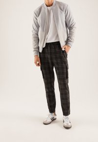 Chaqueta bomber gris con mangas en un gris claro contrastante, camiseta blanca, pantalones cargo de cuadros negros, y zapatillas grises con rayas blancas.
