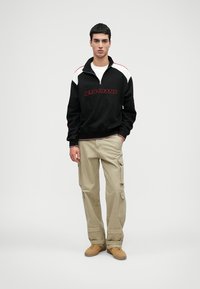 Μαύρο φούτερ με φερμουάρ και κόκκινο κείμενο, με λευκές διακοσμήσεις στους ώμους, συνδυασμένο με μπεζ cargo παντελόνι και φθάνες σε ταμπά χρώμα, σε απλό φόντο.