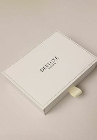Witte rechthoekige doos met een textuurrijke afwerking. Voorzien van een zwart logo met de tekst "DEELUXE WOMEN" en een klein stoffen lipje om te openen.