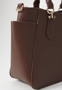 Sac à main en cuir texturé marron avec quincaillerie dorée, comportant une poche latérale et une forme structurée. Fini lisse et poignée robuste.